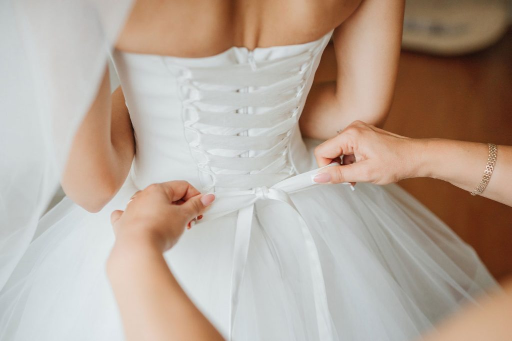 Pessoa vestida de noiva 2023 com corpete enquanto outra pessoa ajusta um laço nas costas.